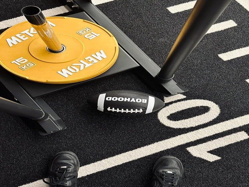 Athletes in a gym performing sled pushes and agility ladder drills on a durable green padded turf surface.